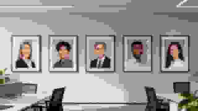 Five framed headshots of diverse professionals displayed on an office wall above empty desks.