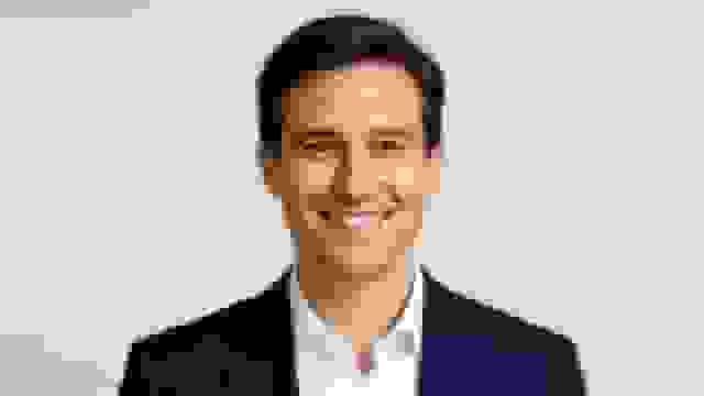 A professional headshot of a smiling man in a dark blue suit and white shirt.